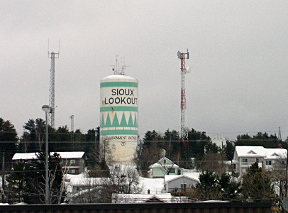 [The white cylindrical tower with a light green design has Sious Lookout near the top. There is an antenna tower on either side of it and other smaller buildings around it all with snow on them.]